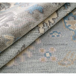 May include: Close-up of a light blue rug with a geometric pattern in shades of beige, light blue, and gray. The rug is folded in half, showing the texture of the fabric.