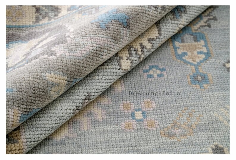 May include: Close-up of a light blue rug with a geometric pattern in shades of beige, light blue, and gray. The rug is folded in half, showing the texture of the fabric.