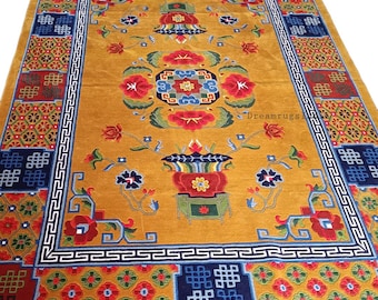 May include: A traditional Tibetan rug with a gold background and intricate floral patterns. The rug features a central design with a vase and flowers, surrounded by a border of geometric patterns and knots. The rug is made of wool and has a soft, luxurious feel.