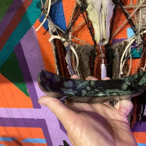 May include: A dark, oblong, polished stone bowl with green and black patterns. The bowl is held in a hand, with a colorful geometric background and a decorative wall hanging with a skull.