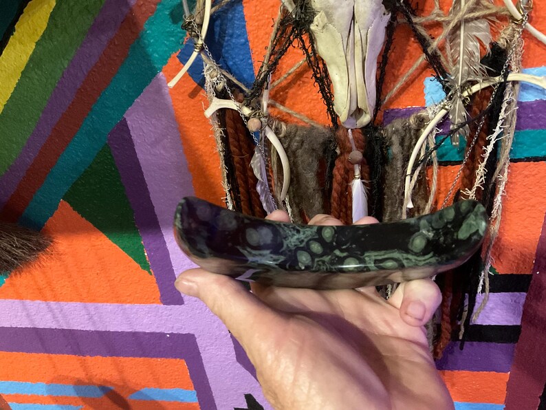 May include: A dark, oblong, polished stone bowl with green and black patterns. The bowl is held in a hand, with a colorful geometric background and a decorative wall hanging with a skull.