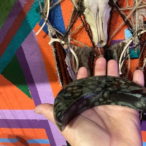 May include: A dark green, patterned stone crescent held in a hand. The background features a colorful geometric design with orange, purple, and blue stripes, and a decorative wall hanging with a skull and feathers.