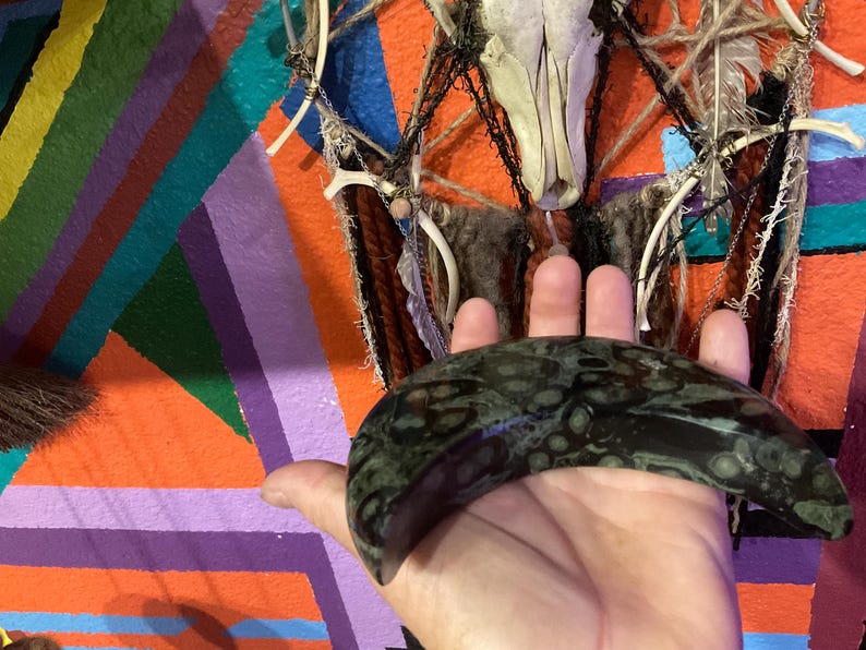 May include: A dark green, patterned stone crescent held in a hand. The background features a colorful geometric design with orange, purple, and blue stripes, and a decorative wall hanging with a skull and feathers.