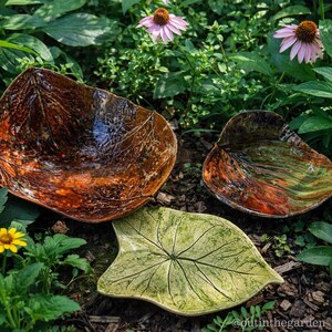May include: Three ceramic leaf-shaped bowls in shades of brown, green, and yellow. The bowls are placed among green plants and flowers. The bowls have detailed vein patterns and a glossy finish, perfect for serving or decoration.