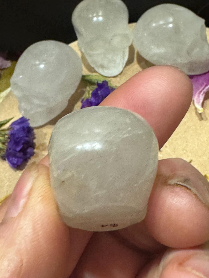 May include: A collection of small, translucent quartz crystal skull figurines. One skull is held between fingers, with others arranged nearby. The skulls are carved with detailed features and have a polished appearance. Purple flowers are scattered around the skulls.