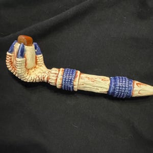 May include: A ceramic pipe featuring a textured, bone-colored handle shaped like a bird claw clutching a smooth, amber-colored stone. The design includes two wide, dark blue bands with a raised, dotted texture encircling the elongated stem.