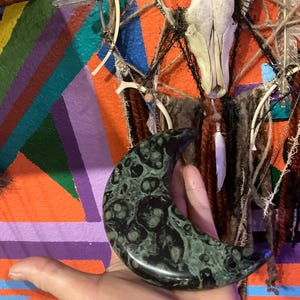 May include: A crescent-shaped, dark green stone with a mottled pattern is held in a hand. The background features a colorful, geometric painted design with orange, purple, and teal stripes, and a decorative wall hanging.