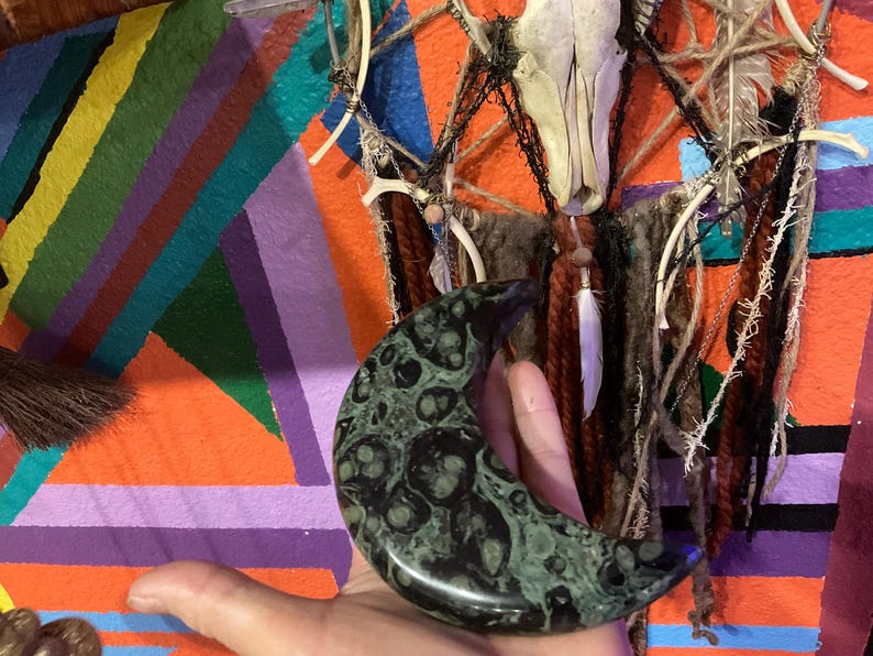 May include: A crescent-shaped, dark green stone with a mottled pattern is held in a hand. The background features a colorful, geometric painted design with orange, purple, and teal stripes, and a decorative wall hanging.