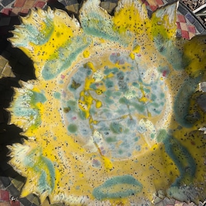 May include: A round ceramic bowl with a scalloped, petal-like edge. The glaze features a vibrant mix of bright yellow, soft teal, and white, with dark speckles scattered across the surface, creating a textured, multi-colored appearance.