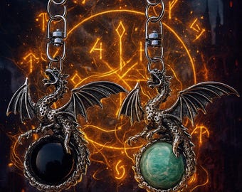 May include: Two silver dragon keychains. Each dragon has outstretched wings and is perched atop a gemstone. One gemstone is black, the other is green. The background features an orange glowing design.
