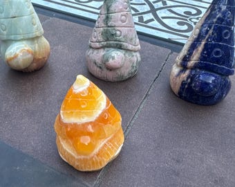 May include: Four carved stone gnome figurines in shades of green, pink, blue, and orange. Each gnome has a pointed hat and a round body. The figurines are displayed on a dark surface.