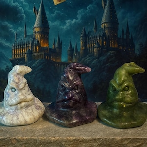 Könnte beinhalten: Drei Sortierhüte aus Stein, jeder mit einer einzigartigen Farbe und Textur. Die Hüte sind vor der Kulisse eines Schlosses unter einem mondbeschienenen Himmel mit einer Eule, die einen Brief trägt, platziert. Die Hüte sind weiß, lila und grün.