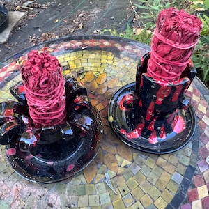 May include: Two black ceramic holders with red drip glaze patterns, each containing a thick, cylindrical bundle of dried red sage leaves tightly wrapped with red twine, positioned upright on a circular base with curved decorative side accents.