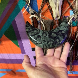 May include: A crescent-shaped, polished stone with a dark green and black mottled pattern. The stone is held in a hand against a colorful, abstract background with orange, purple, and green hues. The stone's shape resembles a stylized moon.