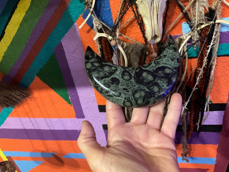 May include: A crescent-shaped, polished stone with a dark green and black mottled pattern. The stone is held in a hand against a colorful, abstract background with orange, purple, and green hues. The stone's shape resembles a stylized moon.
