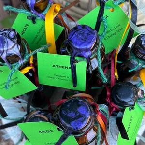 May include: A collection of small glass bottles with cork stoppers sealed with marbled purple and black wax. Each bottle features a bright green rectangular tag with printed text including names like Hecate, Athena, and Brigid, tied with colorful ribbons.