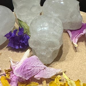 May include: Four clear quartz crystal skull figurines. The skulls are surrounded by dried purple, pink, and yellow flower petals. The skulls are on a light brown surface.