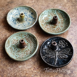 May include: Four ceramic incense holders with central cones. The holders feature floral and dragonfly designs in shades of blue, green, and black. The bottom right holder has the text "TattooPottery".