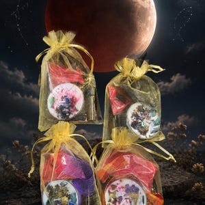 May include: Four small round candles topped with dried flowers and crystals, each individually packaged in a sheer gold organza drawstring bag. The candles feature white wax bases decorated with colorful botanical elements and small stones.