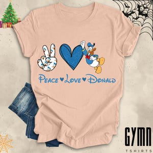 Pode incluir: Uma camiseta cor de p&ecirc;ssego com um sinal de paz, um cora&ccedil;&atilde;o e um gr&aacute;fico do Donald Duck. O texto "Peace Love Donald" &eacute; impresso abaixo do gr&aacute;fico.