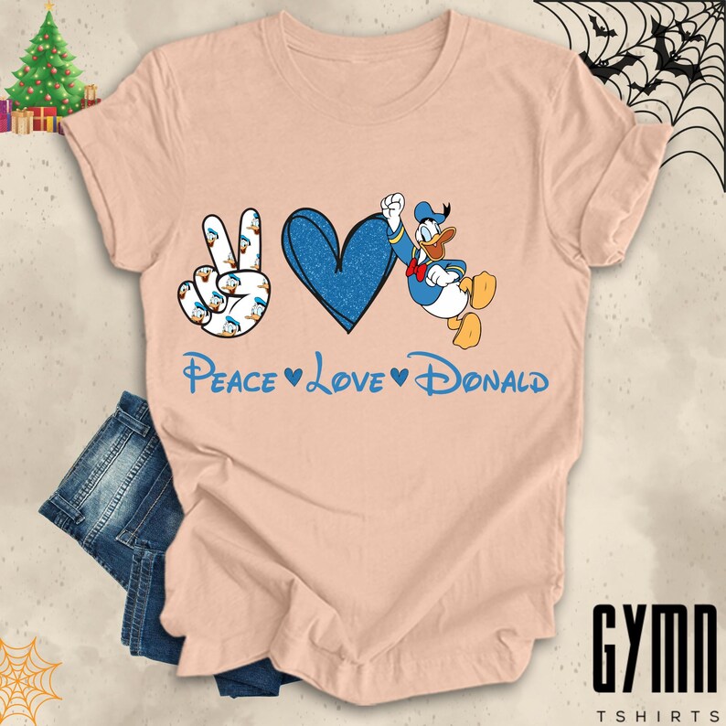 Pode incluir: Uma camiseta cor de p&ecirc;ssego com um sinal de paz, um cora&ccedil;&atilde;o e um gr&aacute;fico do Donald Duck. O texto "Peace Love Donald" &eacute; impresso abaixo do gr&aacute;fico.