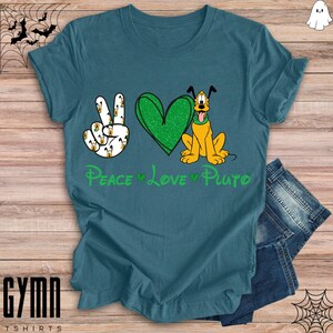 Pode incluir: Uma camiseta azul turquesa com um s&iacute;mbolo de paz verde, um cora&ccedil;&atilde;o verde e uma imagem de desenho animado do Pluto. O texto "Peace Love Pluto" est&aacute; impresso na camiseta.