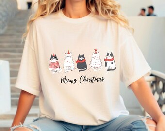 May include: A white t-shirt featuring a row of cartoon cats with festive accessories. The cats are wearing hats and scarves, with the text "Meowy Christmas" below in a handwritten style. The shirt is a casual, holiday-themed design.
