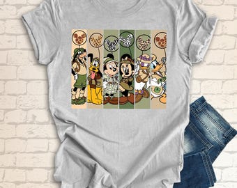 May include: A gray t-shirt with a colorful graphic print of Mickey Mouse, Minnie Mouse, Goofy, Donald Duck, and Pluto dressed in safari outfits. The characters are standing in front of a striped background with animal print patterns.