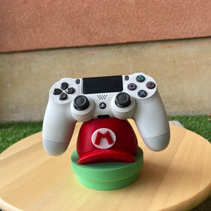 Pode incluir: Um controlador branco do PlayStation 4 está em pé sobre um suporte em forma de chapéu de Mario vermelho e verde. O suporte está sobre uma superfície de madeira.