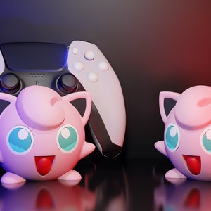 May include: A white and black PlayStation 5 controller is positioned behind two pink Jigglypuff figures. The figures have blue eyes, red tongues, and black-tipped ears. The background is dark with red and blue lighting.
