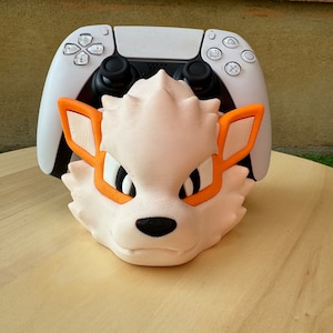 Puede incluir: Una cabeza de animal impresa en 3D de color blanco y naranja, posiblemente un zorro, está sentada encima de un mando de PlayStation 5 blanco.