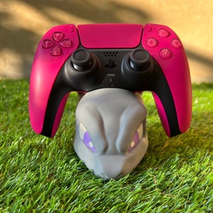 Op de afbeelding: Een roze PlayStation 5 DualSense-controller staat op een grijze 3D-geprinte standaard in de vorm van een Pokémon. De standaard heeft paarse ogen en een grijze neus.
