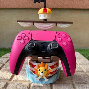 Może przedstawiać: Różowy kontroler PlayStation 5 jest zamontowany na stojaku wydrukowanym w 3D w kształcie statku pirackiego. Statek ma biały żagiel, brązowy maszt i czarną flagę. Figurą dziobową statku jest postać z kreskówki z pomarańczowymi włosami i białym zarostem. Stojak stoi na szarej powierzchni wyłożonej płytkami.