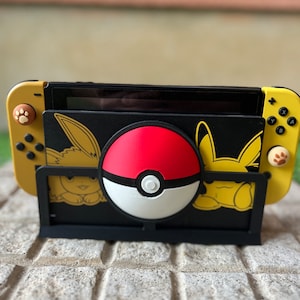 Puede incluir: Un soporte negro con una consola Nintendo Switch amarilla y negra y una Poké Ball roja y blanca. La consola tiene un diseño de Pikachu y Eevee.