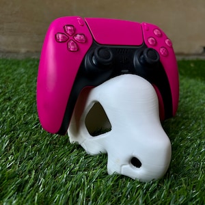 Può includere: Un controller PlayStation 5 rosa con dettagli neri è posizionato su un teschio bianco stampato in 3D. Il teschio ha un foro nella parte anteriore ed è posizionato sull'erba verde.