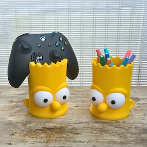 Könnte beinhalten: Zwei gelbe Schreibtisch-Organizer im Bart Simpson-Design. Einer hält einen schwarzen Xbox-Controller, der andere Stifte und Bleistifte. Die Organizer haben Barts Gesicht mit großen Augen und einer kleinen Nase. Der Hintergrund ist eine Holzoberfläche.
