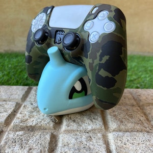 Può includere: Un controller PlayStation 5 con motivo mimetico è montato su un supporto in plastica blu chiaro a forma di creatura dei cartoni animati.