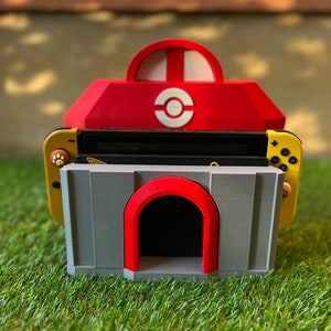 Può includere: Un supporto per Nintendo Switch rosso e bianco con una base grigia e un arco rosso. Il supporto ha una console Switch gialla con pulsanti a forma di zampa. Il design è ispirato a un videogioco popolare.