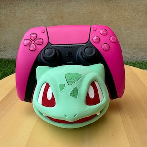Może przedstawiać: Różowy kontroler PlayStation 5 z zielonym stojakiem w kształcie postaci Pokemon Bulbasaur. Kontroler ma czarną przednią płytę z logo PlayStation i przyciskami.