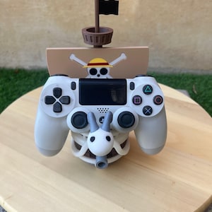 Może przedstawiać: Biały kontroler PlayStation 4 umieszczony jest w stojaku wydrukowanym w 3D w kształcie statku pirackiego. Statek ma brązowy kadłub, czarną flagę i wzór czaszki i skrzyżowanych kości z słomkowym kapeluszem na czaszce. Stojak znajduje się na drewnianej powierzchni.