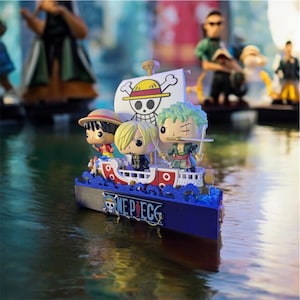 Op de afbeelding: Een miniatuur speelgoedboot met personages uit de One Piece anime serie. De boot is blauw, rood en wit, met een Jolly Roger vlag. De personages Luffy, Sanji en Zoro zijn te zien. De boot heeft de tekst "ONE PIECE" op de voorkant.
