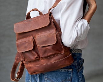 Puede incluir: Una mochila de cuero marrón con dos bolsillos grandes en la parte delantera y un cierre de solapa. La mochila la lleva puesta una persona que lleva una camisa blanca y unos vaqueros azules.