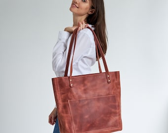 Puede incluir: Un bolso tote de cuero marrón con un bolsillo frontal. El bolso tiene una correa larga y lo lleva una persona.