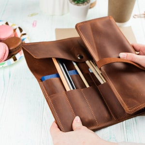 Puede incluir: Un rollo de l&aacute;pices de cuero marr&oacute;n con m&uacute;ltiples ranuras para guardar l&aacute;pices, bol&iacute;grafos y otros materiales de arte. El rollo est&aacute; abierto y las ranuras son visibles.