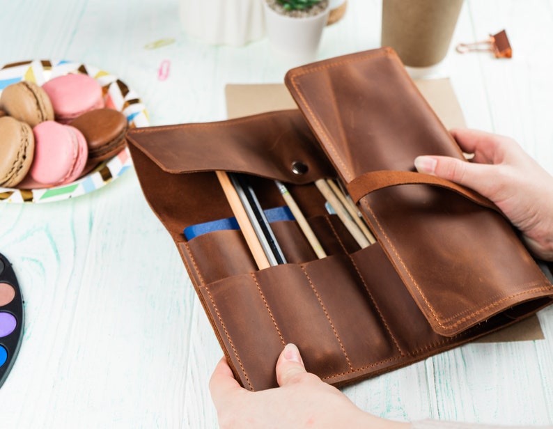 Puede incluir: Un rollo de l&aacute;pices de cuero marr&oacute;n con m&uacute;ltiples ranuras para guardar l&aacute;pices, bol&iacute;grafos y otros materiales de arte. El rollo est&aacute; abierto y las ranuras son visibles.