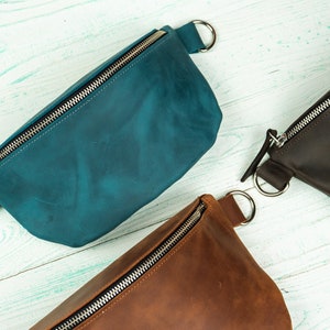 K&ouml;nnte beinhalten: Leder-Bauchtaschen in Teal, Braun und Dunkelbraun. Jede Tasche hat einen silbernen Rei&szlig;verschluss und einen silbernen Ring auf jeder Seite. Die Taschen sind auf einer hellblauen Holzoberfl&auml;che angeordnet.