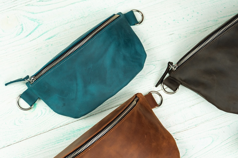 K&ouml;nnte beinhalten: Leder-Bauchtaschen in Teal, Braun und Dunkelbraun. Jede Tasche hat einen silbernen Rei&szlig;verschluss und einen silbernen Ring auf jeder Seite. Die Taschen sind auf einer hellblauen Holzoberfl&auml;che angeordnet.