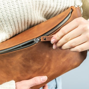 K&ouml;nnte beinhalten: Eine braune Leder-Bauchtasche mit silbernem Rei&szlig;verschluss. Die Tasche ist rechteckig mit einer geschwungenen Form und einem kleinen Leder-Rei&szlig;verschluss-Anh&auml;nger. Die Tasche wird ge&ouml;ffnet.