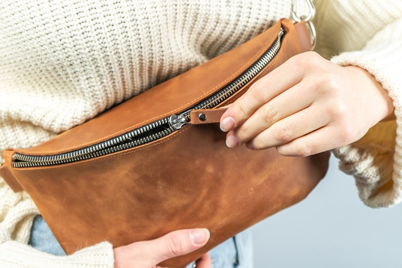 K&ouml;nnte beinhalten: Eine braune Leder-Bauchtasche mit silbernem Rei&szlig;verschluss. Die Tasche ist rechteckig mit einer geschwungenen Form und einem kleinen Leder-Rei&szlig;verschluss-Anh&auml;nger. Die Tasche wird ge&ouml;ffnet.
