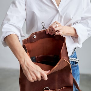 Puede incluir: Una mochila de cuero marr&oacute;n con cierre de solapa, abierta para revelar el interior. La bolsa tiene un cierre plateado y un asa peque&ntilde;a. El interior presenta bolsillos y una correa negra. La persona lleva una camisa blanca y vaqueros azules.